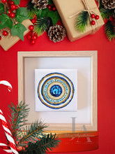 A colorful mandala-style original artwork in a wooden frame , blue , gold glitter pattern, surrounded by Christmas decorations including pine branches, holly , and a pine cone, on red background