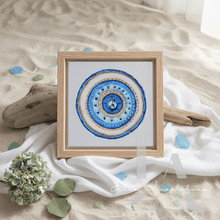 A square framed original art featuring a circular mandala design in shades of blue, gold, and white, styled on a white draped cloth over a sandy surface with driftwood and green hydrangea flowers.