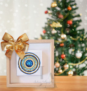 A framed decorative wall art original painting, in mandala-style, with a blue and gold glitter design, placed on a wooden surface in front of a Christmas tree with lights and ornaments. Perfect art gift for someone special. The canvas painting, with greek evil eye, is 4x4 inch, and with the frame 8x8.