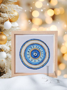 A framed original greek evil wall art in circular mandala design with intricate patterns and colors, including shades of blue , white , and gold glitter, set against a blurred background with Christmas decorations.