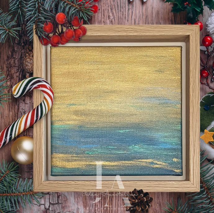 A framed original painting depicting a serene abstract ocean minimalist landscape with gold and blue color scheme. The frame is surrounded by Christmas decorations such as pine branches, red berries, and a candy cane.