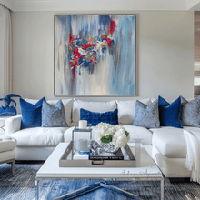 A large square abstract canvas painting with blue, white, dark berry pink, and silver gray brushstrokes hangs on a beige wall above a white sectional sofa styled with blue and gray pillows in a modern living room.