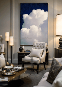 A beige armchair with a pillow in a living room with a large painting of white clouds and celestial night blue sky in the background. The room features decorative elements like two lamps and a black low coffee table. The overall atmosphere is calm and serene
A large painting of a white cloud against a dark blue celestial sky hangs above a wooden shelf in a minimalist living room . The room features decorative elements like vases with flowers , and wooden low coffee table . The overall atmosphere is calm and