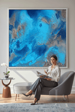 A woman sits in a gray lounge chair in a modern living room, holding a magazine in front of a large framed abstract painting with swirling blue, teal, and silver tones.