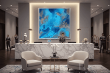 A large square abstract painting with vibrant blue, teal, and silver marbled patterns hangs on a dark gray wall above a modern white marble reception desk in a luxury hotel lobby. Two cream-colored armchairs sit in the foreground on a patterned rug.