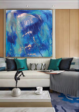 A large abstract canvas painting with vibrant blue, teal, and white brushstrokes hangs on a light wood-paneled wall above a modern white sectional sofa with teal and gray pillows.