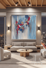 A large framed abstract painting with blue, white, dark pink, and gray brushstrokes hangs on a gray wall above a light beige sofa in a modern living room with city views at night.