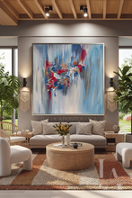 A large framed abstract painting with blue, white, dark berry pink, and gray brushstrokes hangs on a gray wall above a light beige sofa in a modern living room with plants, a round woven coffee table, and warm ambient lighting.
