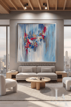 A large framed abstract canvas painting featuring expressive brushstrokes in blue, white, dark berry pink, gold and silver, hangs on a gray wall above a modern light gray sofa in a bright, contemporary living room with city views.