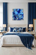 An abstract canvas wall art print featuring vibrant shades of blue and navy with silver highlights, displayed in a modern bedroom setting above a neatly made bed with matching blue and white bedding.