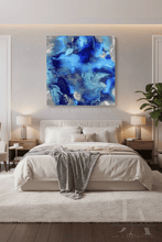 A modern bedroom featuring a square abstract canvas print with vibrant blue and navy fluid art hanging above a neatly made bed. The room is styled with a white shag rug, wooden nightstands, matching lamps, and a green plant on the right side.
