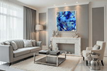 A modern living room featuring a square abstract blue and silver canvas print hanging above a white marble fireplace mantel. The room is styled with a light gray sofa, a white armchair, a glass coffee table, and a white shag rug.