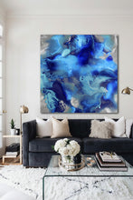 A large square abstract canvas print with swirling luminous blue, navy, silver, and teal fluid patterns hangs on a light wall above a dark gray sofa in a modern living room setting.