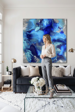 A woman stands in a modern living room in front of a large abstract canvas print featuring swirling shades of blue, navy, and silver. The room includes a dark gray sofa with neutral throw pillows, a glass coffee table with white flowers, and a white shag rug.