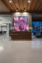A large, vibrant abstract painting with swirling purple, pink, and blue hues is displayed on a wall in a modern, upscale lobby. The painting is framed in gold and hangs above a dark wood reception desk decorated with white flowers.