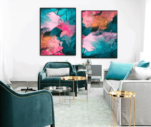 A modern living room featuring two tall abstract canvas prints with vibrant teal, pink, and orange brushstrokes, hung on a white wall above a teal armchair and a light gray sofa.