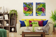 A pair of colorful abstract canvas wall art prints hanging above a beige sofa in a styled living room. The canvases feature vibrant brushstrokes of blue, green, yellow, and pink, and the room is decorated with a wooden coffee table, colorful pillows, and plants.