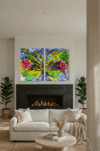 A modern living room featuring a two-panel canvas wall art set depicting a colorful abstract floral scene with blue, green, yellow, and pink brushstrokes, hung above a black fireplace. The room is styled with a white sofa, potted green plants, and a round wooden coffee table.