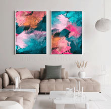 Two vertical framed abstract canvas prints with vibrant pink, teal, orange, and black brushstrokes, displayed side-by-side on a white wall above a beige sectional sofa in a modern living room setting.
