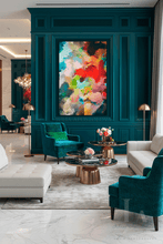 A luxury modern living room featuring a large, vibrant abstract canvas painting with red, yellow, green, and blue colors, mounted on a deep teal wood-paneled wall. The room is furnished with matching teal velvet armchairs, white leather sofas, gold floor lamps, and round coffee tables on a light gray rug.
