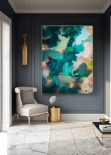 A large framed abstract canvas painting with swirling teal, green, blue, pink, and gold tones hangs on a dark charcoal wall in a modern styled room with a cream armchair, gold side table, marble floor, and patterned rug.