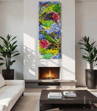 A modern living room with a white sofa, dark wood coffee table, potted plants, and a fireplace, featuring two vertical abstract canvas prints hanging above the fireplace. The canvases contain vibrant green, blue, yellow, and pink brushstrokes.