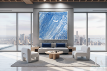 A large square abstract blue painting with white and light gray brushstrokes hangs on a gray wall above a modern light gray sofa in a bright, contemporary living room with city views through floor-to-ceiling windows.