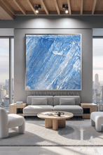 A large square abstract painting with diagonal cool blue and white brushstrokes hangs on a gray wall above a modern light gray sofa in a bright, contemporary living room with city views.