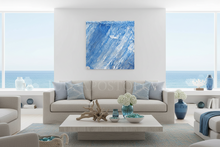 A square abstract canvas print with blue and white brushstrokes hangs on a white wall above a modern beige sofa in a bright, contemporary living room with a view of the ocean.