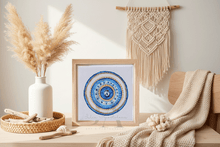 Framed mandala art piece on a wooden surface with decorative elements like a vase, macrame wall hanging, and dried plants.