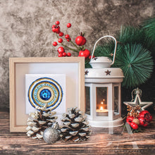 A white lantern with a lit candle inside, surrounded by pine branches , red berries , pine cones , and a framed original glitter artwork with a colorful mandala design on a wooden table