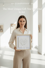 Woman holding a framed artwork of white heart in a bright room with light streaming through windows.