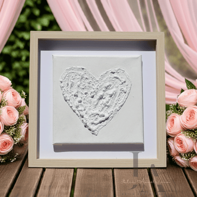 Framed heart-shaped artwork on a wooden surface with pink roses and curtains in the background