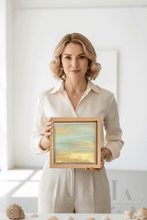 A woman with blonde hair wearing a cream-colored blouse holds a small original framed painting depicting an minimalist abstract beach scene with turquoise water and golden sand. The background is a bright, minimalist white room with a table in the foreground decorated with seashells.