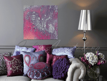 An abstract canvas wall art print with pink, purple, and silver tones, displayed in a styled living room setting above a sofa with decorative pillows and next to a silver floor lamp.