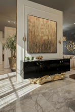 A large abstract wall art piece with textured gold and bronze tones hangs on a white architectural wall above a glossy black console table in a modern, sunlit interior with marble flooring.