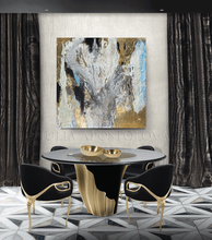 Modern dining room with large gold leaf  abstract painting on the wall, a round table and black chairs with gold, velvet curtains, and geometric floor pattern.