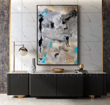 A framed abstract canvas print with gray, black, gold, and turquoise paint strokes, displayed on a marble-tiled wall above a modern black console table with gold accents.