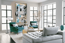 A modern living room featuring a large vertical abstract canvas print with black, white, gray, and blue tones hanging on a light gray wall. The room is furnished with two teal velvet armchairs, a light gray tufted chaise lounge, a light wood coffee table, and a tall white vase with dried branches.