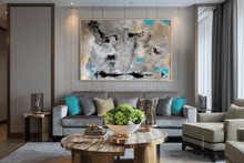 A large framed abstract canvas painting with gold, gray, black, and turquoise accents hangs on a gray wall above a modern gray sofa in a styled living room.