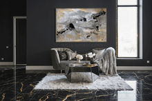 An abstract canvas painting with black, white, and gold brushstrokes hangs on a dark charcoal wall above a gray tufted sofa in a modern living room setting with black marble floors.