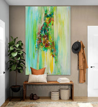 A large vertical abstract canvas painting with vibrant green, yellow, and brown brushstrokes hangs on a taupe entryway wall above a wooden bench with decorative pillows, a potted plant, and a hanging coat.