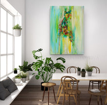 A vertical abstract canvas print with vibrant green, yellow, and brown brushstrokes hangs on a white wall in a modern, sunlit dining room with wooden chairs and potted plants.