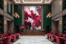 A large abstract wall art piece with swirling burgundy pink, green, purple, and white brushstrokes hangs in a luxurious hotel lobby or lounge. The artwork is framed in gold and set against dark green paneled walls, with pink velvet armchairs, gold chandeliers, and marble flooring visible around it.