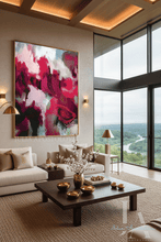 A large framed abstract painting with bold magenta, pink, white, and dark accents hangs on a beige wall in a modern living room with a sofa, coffee table, and large windows overlooking a green landscape.