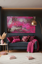 A large rectangular abstract wall art piece with vivid pink, magenta, and metallic gold marbling hangs above a dark blue velvet sofa in a styled living room. The scene includes patterned pillows, a pink throw blanket, a floor lamp, side table, books, and a rug.