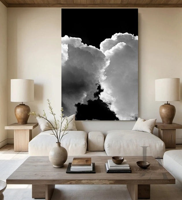 A modern boho living room with a large abstract painting on the wall featuring kissing clouds in shades of white, and gray, against black sky. The room also includes two lamps, a low wooden coffee table, a white couch and earthy neutral colors.