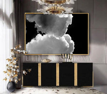 A modern luxury living room with a large framed abstract painting on the wall featuring kissing clouds in shades of white, and gray, against black sky background. The room also includes two vases with gold flowers, gold lighting, over black cadenza furniture with gold accents, creating a luxury atmosphere.