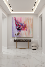 A large abstract canvas painting with vibrant pink, purple, lilac, and peach brushstrokes hangs on a light gray wall in a modern white hallway. Below the painting is a dark wood console table with gold metal legs, and to the right is a small round white and black ottoman.