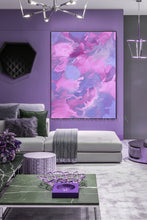 A large vertical abstract canvas print with swirling pink, purple, and lilac paint patterns, displayed on a lavender wall above a modern light gray sofa in a styled living room.